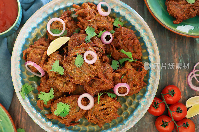 图片:一堆自制的脆洋葱bhajis / pakoras配以香菜叶和红洋葱圈，放在有图案的盘子上，旁边是葡萄藤番茄，印度菜素食小吃，木纹背景，高架视野